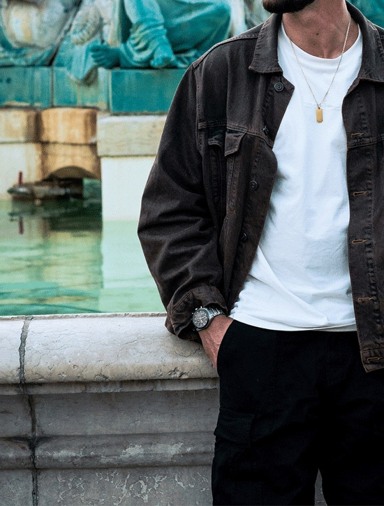 Homme en veste marron et pantalon noir, appuyé sur une structure en pierre