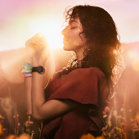 A woman with curly hair, wearing a watch and scrunchie, stands in a field with her eyes closed, bathed in the golden light of the setting sun.
