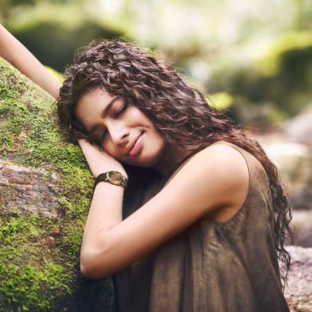 A woman with curly hair rests her head on a mossy tree trunk, eyes closed and a gentle smile on her face, set against a blurred natural background.
