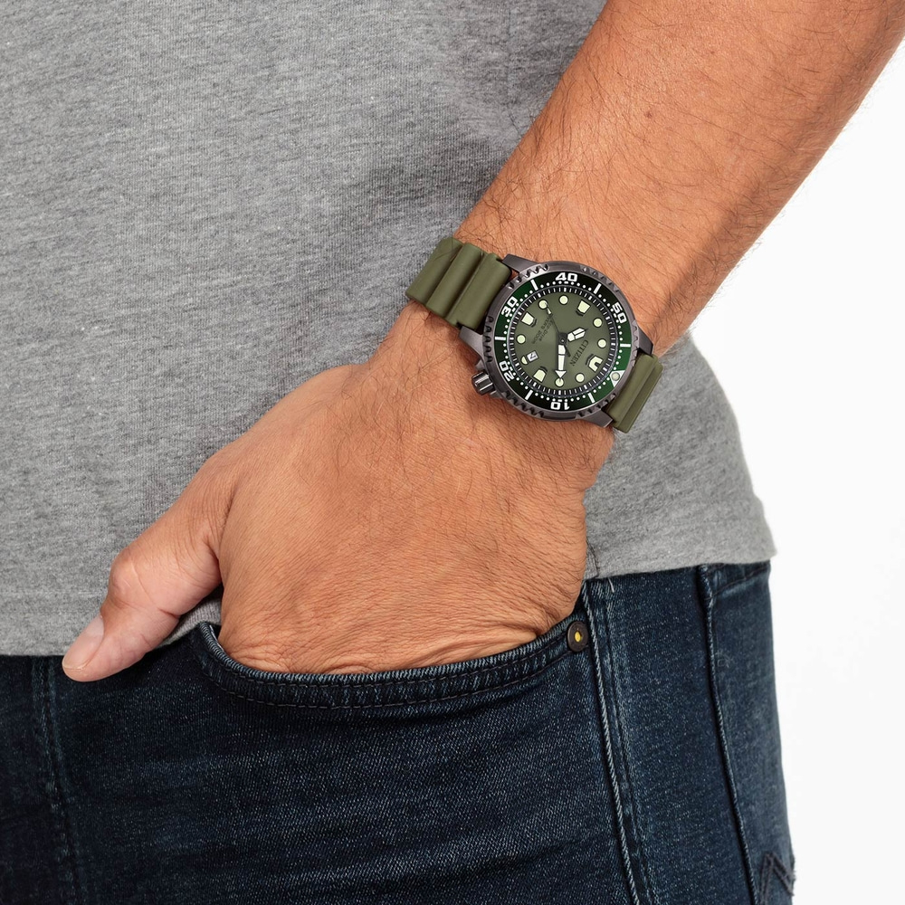 Homme portant une montre verte, main dans la poche, chemise grise, jean bleu.
