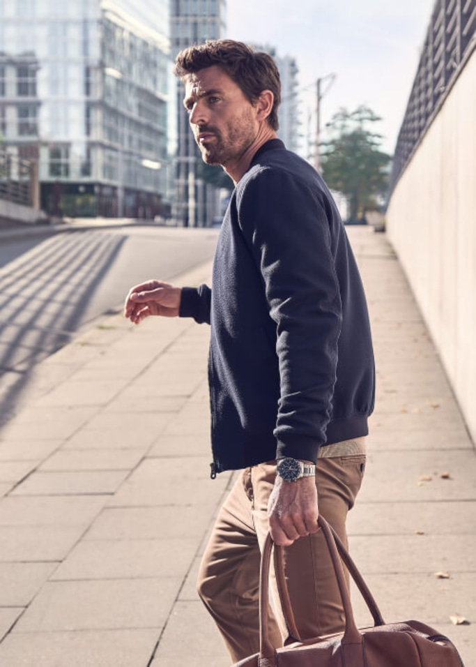 Homme marchant sur le trottoir, tenant un sac, portant une veste et une montre.