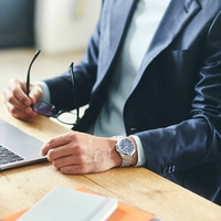 Homme en costume avec montre, tenant des lunettes, à un bureau avec un ordinateur portable.