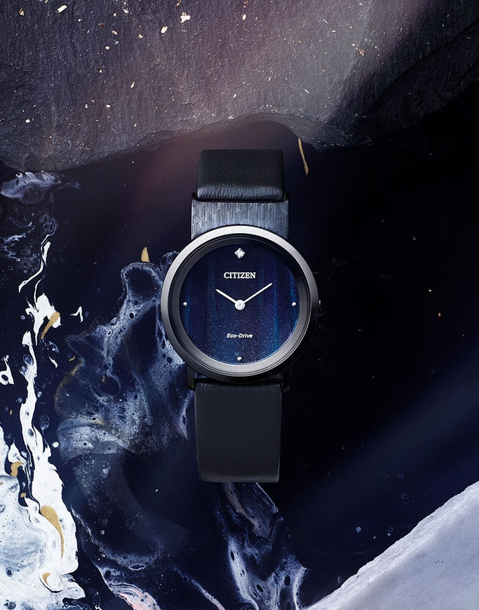 An overhead shot of a Citizen Eco-Drive watch, with a black band and blue galaxy-like face, laying on a dark, textured surface with white and gold swirls.
