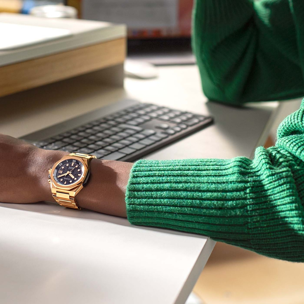 Montre en or au poignet, pull vert, clavier, bureau blanc, environnement de bureau.