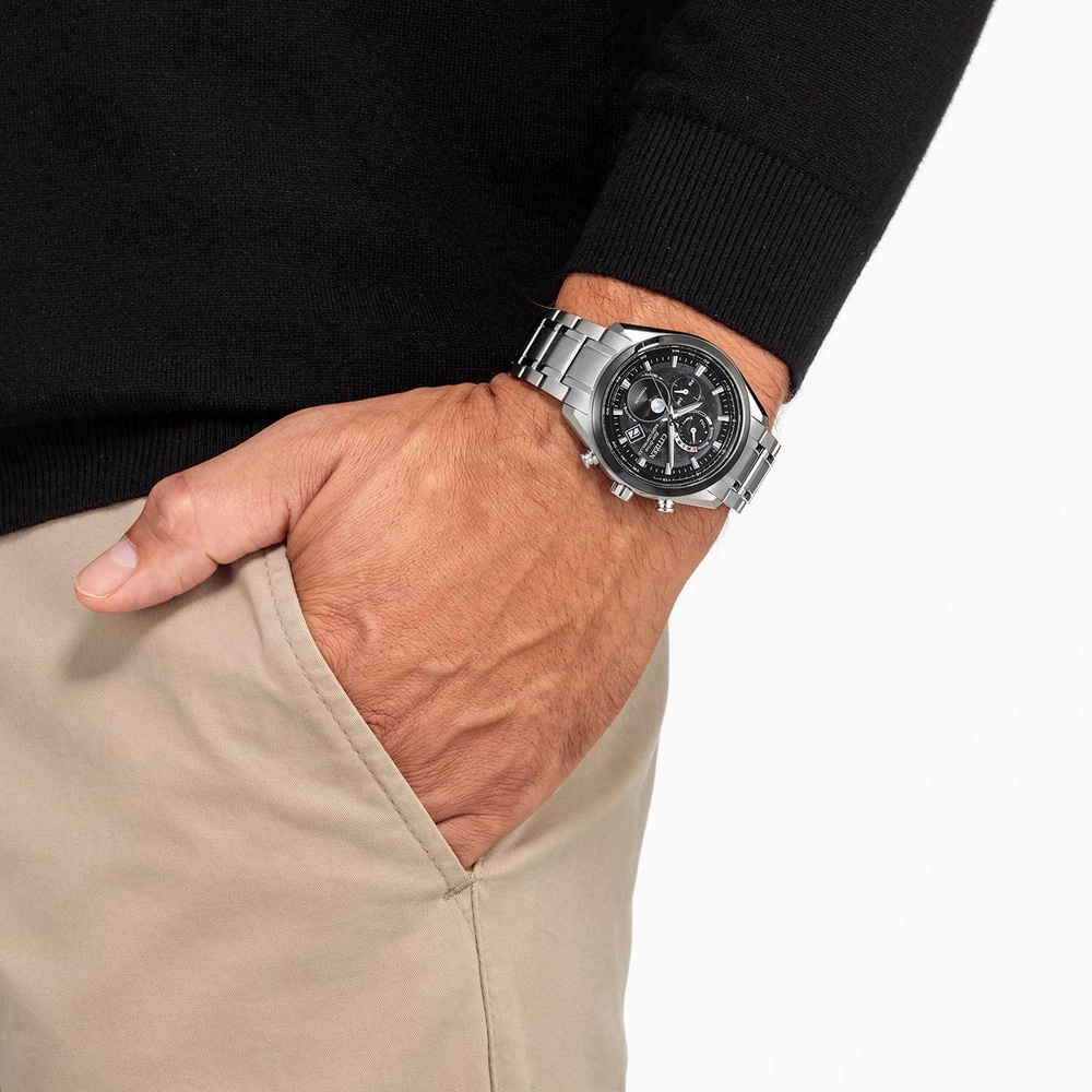 Homme portant une montre, main dans la poche, pull noir, pantalon beige